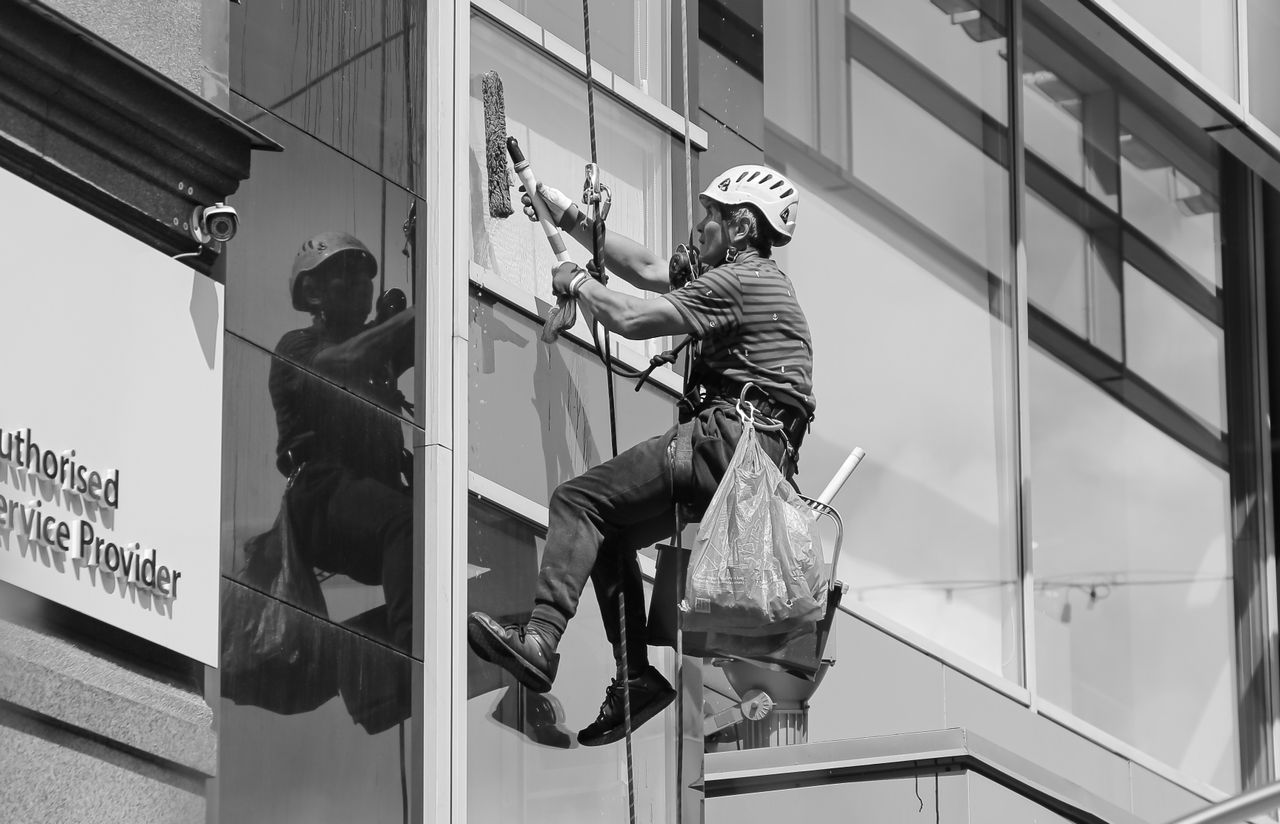 Black and white from below of man steeplejack in protective helmet with professional equipment cleaning glass wall of modern tower